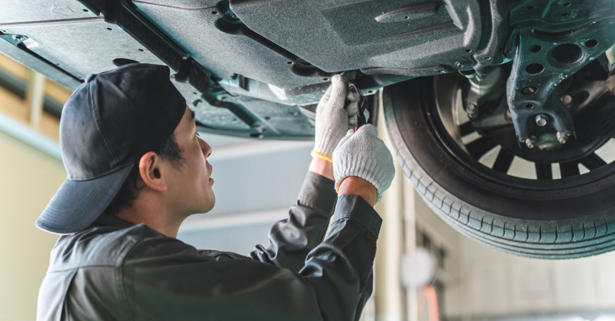 A mechanic wearing blue work clothes reaches into the underbody near the wheel as he works on a lifted vehicle.