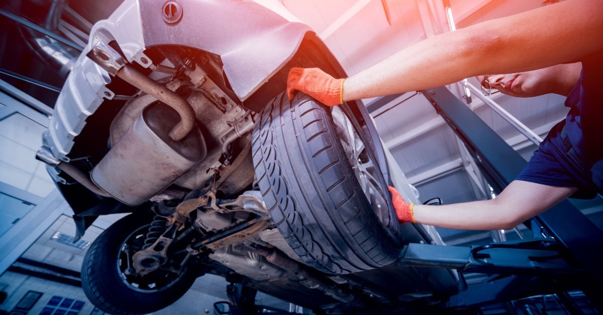 A mechanic grips a rear tire while a lift raises a car, exposing the wheel well, suspension, and undercarriage.