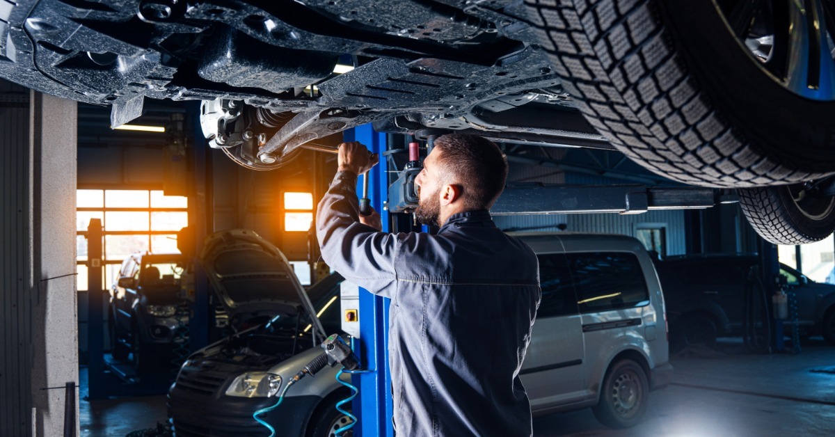 A mechanic works beneath a raised vehicle on a two-post lift while another car sits in the service bay behind him.