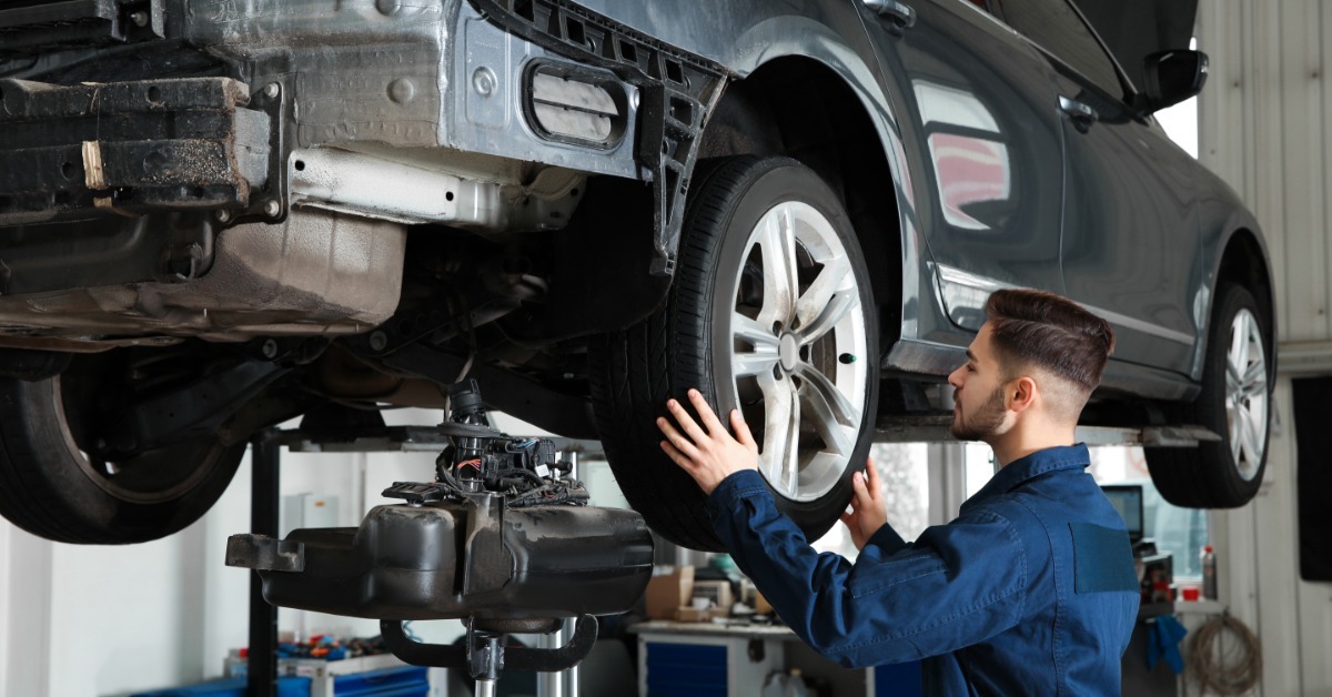 A mechanic checks the front wheel of a raised gray SUV on a two-post lift inside a clean automotive service bay.