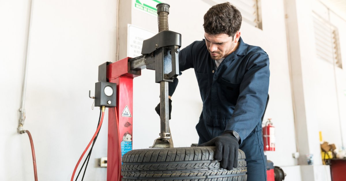 A mechanic uses a tire changer while holding a mounted wheel and tire steady in a bright automotive service bay.