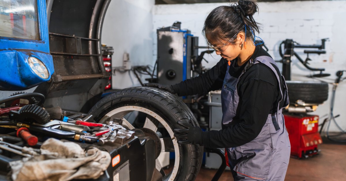 A technician positions a wheel and tire on a wheel balancer while working in a busy automotive repair shop.