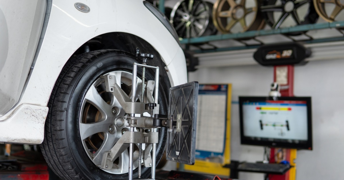 A white car sits on an alignment rack with a wheel alignment sensor attached to its front tire and a diagnostic monitor nearby.